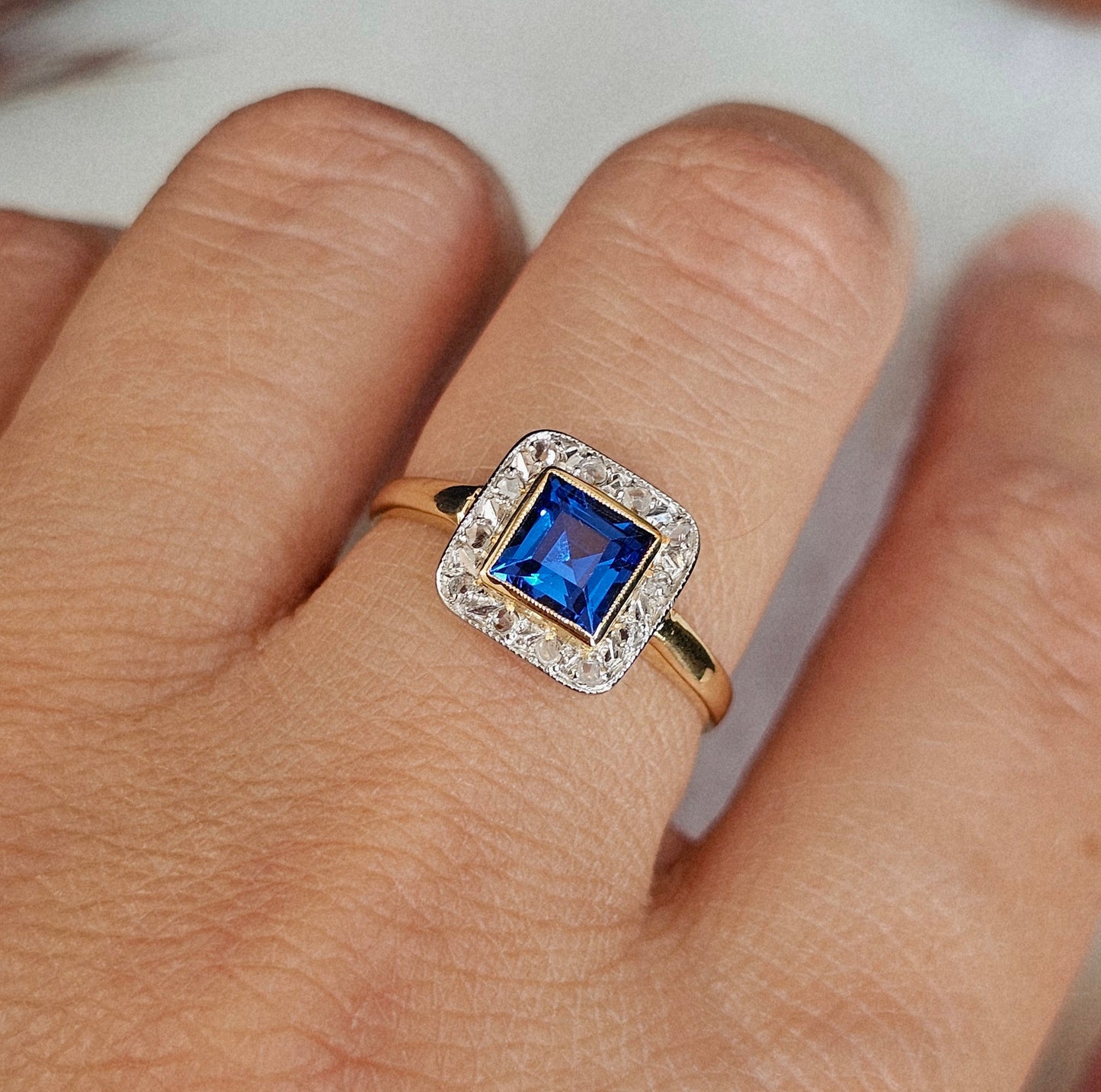 Bague ancienne Art Déco portée, de forme carrée aux coins arrondis. Elle est sertie d'un Spinelle de synthèse bleu intense carré et taillé à degré, entouré d'un pavage de roses de diamant sur Or blanc. La monture est en or jaune 18 carats.